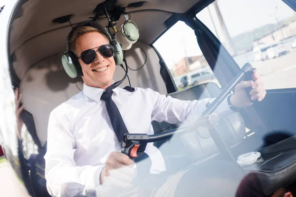 Smiling Pilot in sunglasses and headset with microphone sitting in helicopter cabin and holding wheel — Stock Photo
