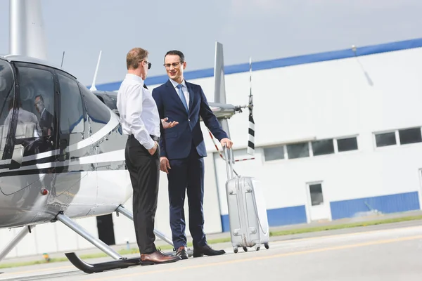 Piloto en ropa formal y hombre de negocios con maleta cerca de helicóptero - foto de stock
