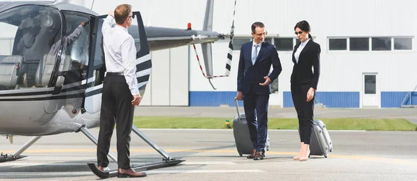 Panoramic shot of business partners with luggage near pilot and helicopter — Stock Photo