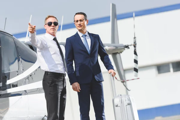 Pilot in formal wear pointing with finger and businessman with suitcase near helicopter — Stock Photo