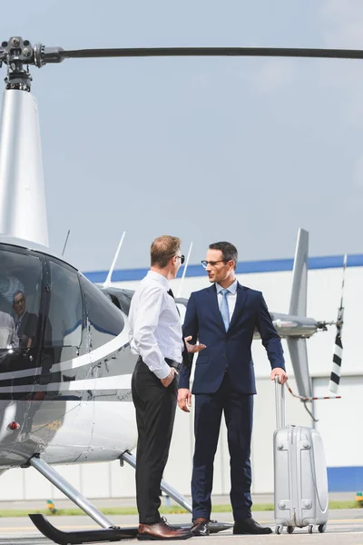 Piloto en ropa formal y hombre de negocios con maleta cerca de helicóptero - foto de stock