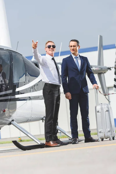 Pilot in offizieller Kleidung zeigt mit Finger und Geschäftsmann mit Gepäck in der Nähe des Hubschraubers — Stockfoto