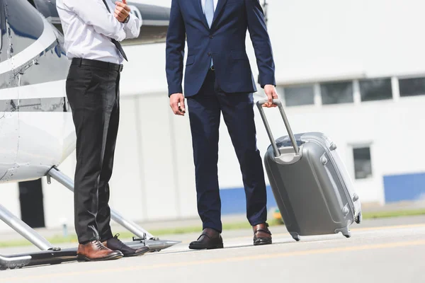 Vista recortada de Piloto en ropa formal y hombre de negocios con equipaje cerca de helicóptero - foto de stock