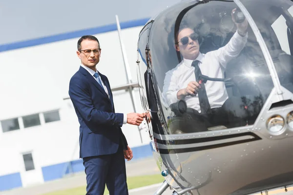 Piloto en ropa formal sentado en helicóptero mientras hombre de negocios mirando a la cámara - foto de stock