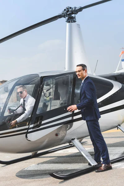 Pilote en tenue formelle assis en hélicoptère pendant que l'homme d'affaires ouvre la porte — Photo de stock