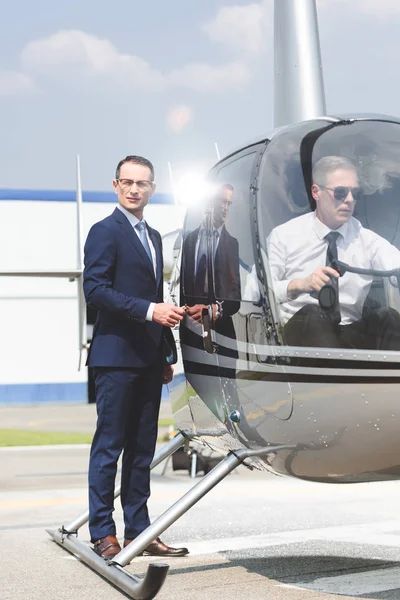 Piloto sentado en helicóptero mientras hombre de negocios en traje abriendo la puerta - foto de stock