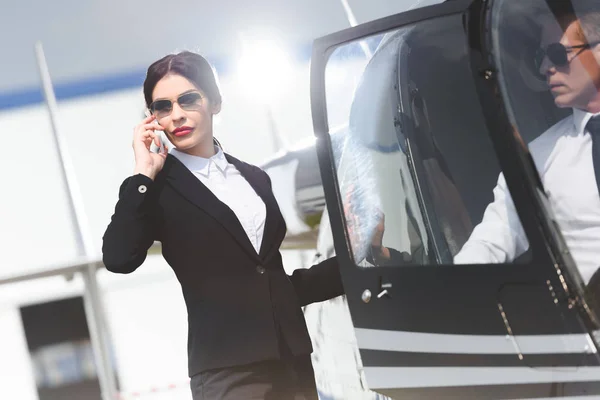 Businesswoman in formal wear talking on smartphone near helicopter with pilot — Stock Photo