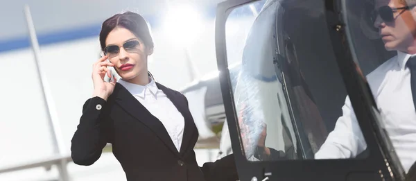 Panoramic shot of businesswoman in formal wear talking on smartphone near helicopter with pilot — Stock Photo