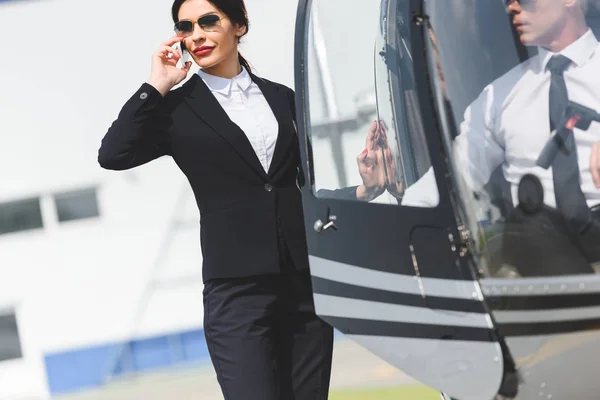 Businesswoman in formal wear talking on smartphone near helicopter with pilot — Stock Photo