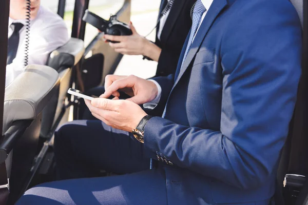 Vista recortada de hombre de negocios en ropa formal sentado en helicóptero con teléfono inteligente - foto de stock