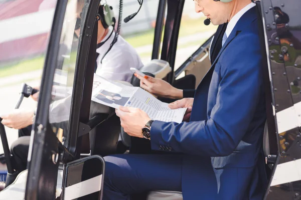 Cropped view of businesspeople in formal wear with newspaper sitting in helicopter with pilot — Stock Photo