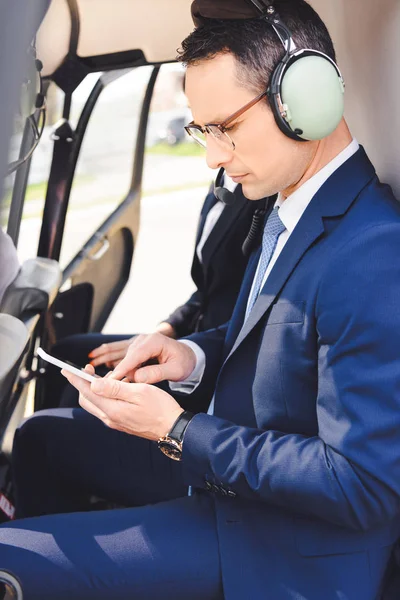 Бизнесмен в формальной одежде и гарнитуре сидит в кабине вертолета и использует смартфон — стоковое фото