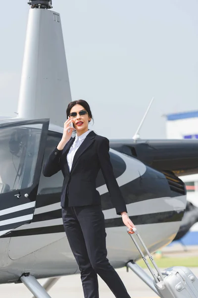 Mujer de negocios en ropa formal con maleta hablando en teléfono inteligente cerca de helicóptero - foto de stock