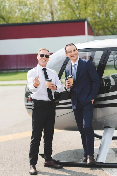 Piloto en gafas de sol mostrando dedo medio y hombre de negocios con café para ir cerca de helicóptero - foto de stock