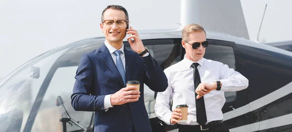 Prise de vue panoramique de l'homme d'affaires avec café pour aller parler sur smartphone tandis que le pilote regardant regarder près de l'hélicoptère — Photo de stock