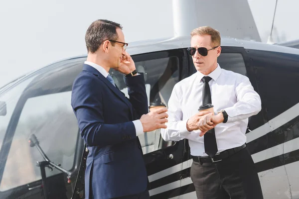 Businessman with coffee to go talking on smartphone while pilot pointing at watch near helicopter — Stock Photo