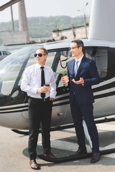 Piloto em óculos de sol e homem de negócios com café para ir perto de helicóptero — Fotografia de Stock