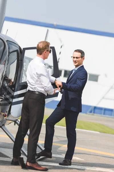 Pilot in offizieller Kleidung in der Nähe von Geschäftsmann öffnet Tür des Hubschraubers — Stockfoto