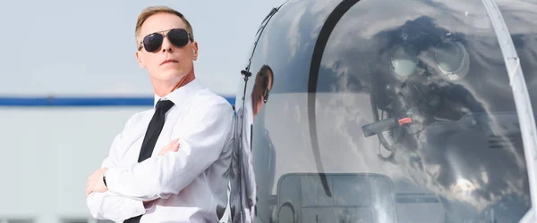 Panoramaaufnahme des Piloten mit Sonnenbrille und formeller Kleidung mit verschränkten Armen in der Nähe des Hubschraubers — Stockfoto