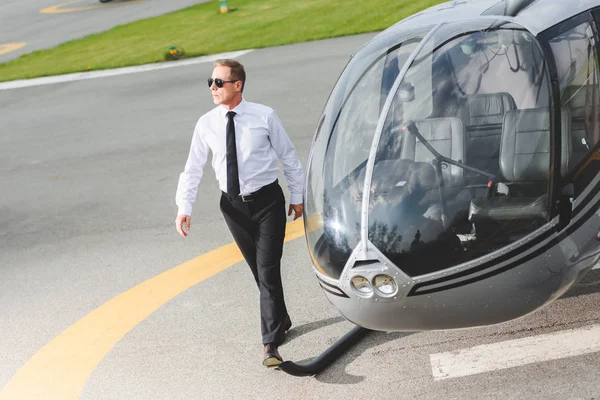 Gut aussehender Pilot in offizieller Kleidung und Sonnenbrille, der in der Nähe des Hubschraubers spaziert — Stockfoto
