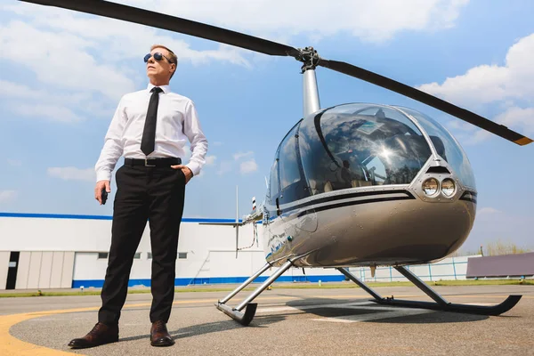 Пілот в офіційному зносу і сонцезахисні окуляри постановки з рук в кишеню біля вертольота — стокове фото