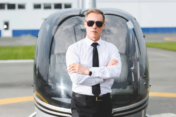 Pilota fiducioso in occhiali da sole e abbigliamento formale con braccia incrociate vicino all'elicottero — Foto stock