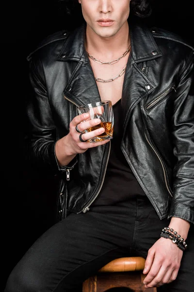 Cropped view of man holding glass of whiskey while sitting on wooden chair isolated on black — Stock Photo