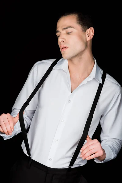 Hombre seguro y guapo en camisa blanca tocando tirantes aislados en negro - foto de stock