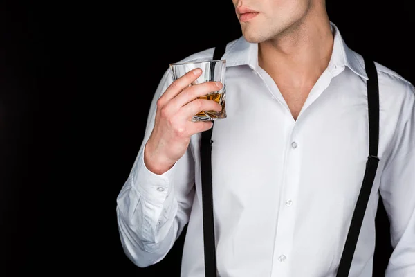 Cropped view of man in white shirt and suspenders holding glass of whiskey isolated on black — Stock Photo