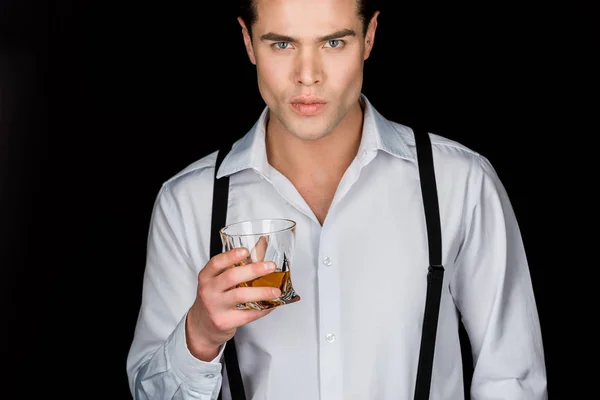Homem bonito em camisa branca e suspensórios segurando vidro de uísque isolado em preto — Fotografia de Stock