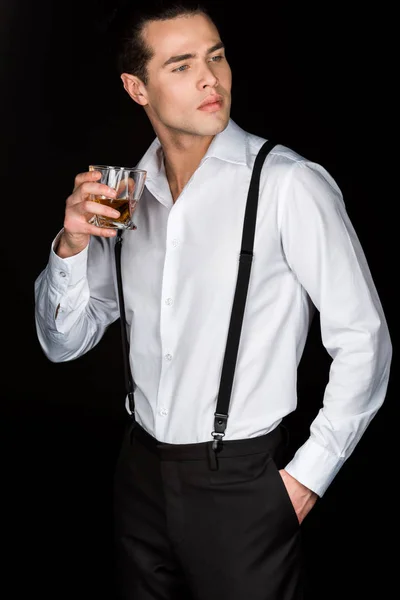 Hombre guapo en camisa blanca y tirantes sosteniendo vaso de whisky mientras está de pie con la mano en el bolsillo aislado en negro - foto de stock