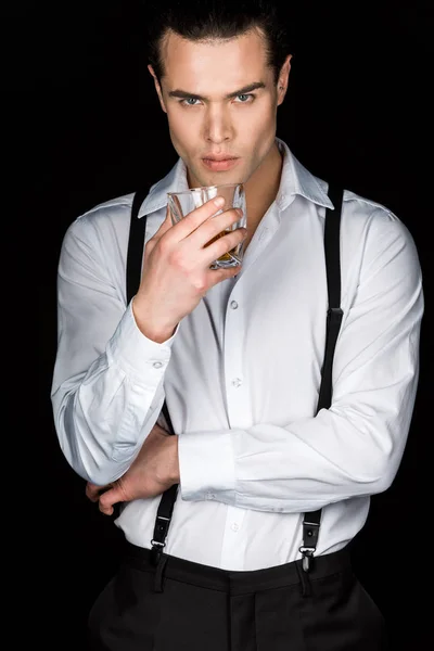 Hombre guapo en camisa blanca y tirantes sosteniendo un vaso de whisky mientras mira la cámara aislada en negro - foto de stock