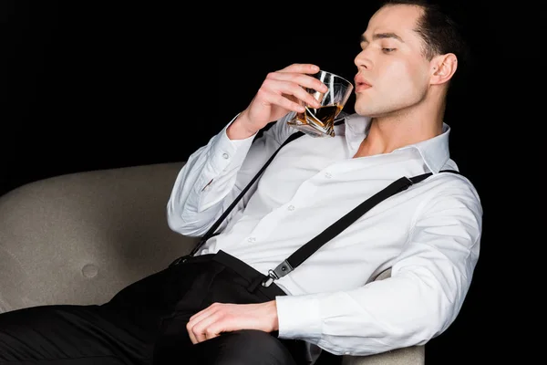 Hombre guapo mirando un vaso de whisky mientras está sentado en sillón aislado en negro - foto de stock