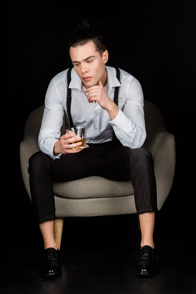 Pensive man in white shirt and suspenders holding glass of whiskey while sitting in armchair isolated on black — Stock Photo
