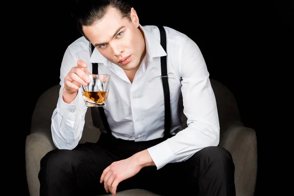 Hombre de camisa blanca y tirantes sosteniendo vaso de whisky mientras está sentado en sillón aislado en negro - foto de stock