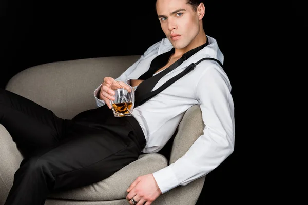 Serious man in white shirt and suspenders holding glass of whiskey while sitting in armchair isolated on black — Stock Photo