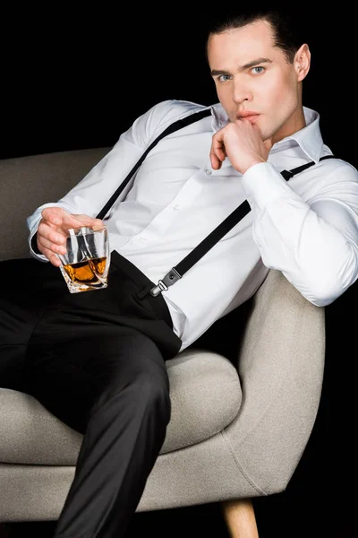 Серьезный человек держит стакан виски и трогательное лицо, сидя в кресле изолированы на черном — стоковое фото