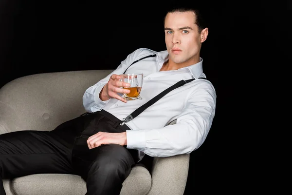 Uomo fiducioso in possesso di un bicchiere di whisky e seduto in poltrona isolato su nero — Foto stock
