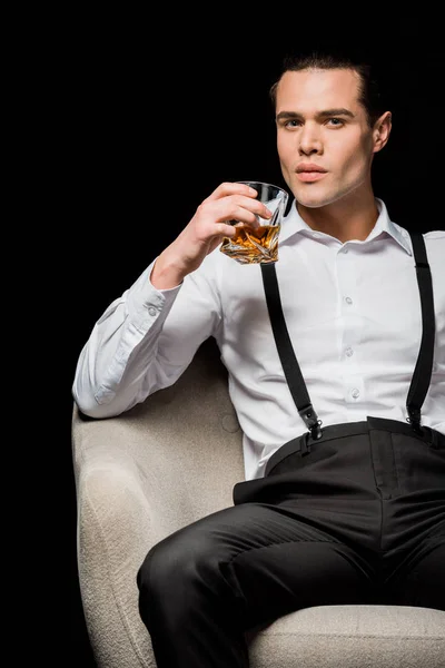 Handsome and confident man in white shirt and suspenders holding glass of whiskey while sitting in armchair isolated on black — Stock Photo