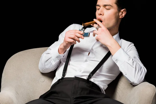 Hombre guapo sosteniendo encendedor cerca de cigarro mientras está sentado en sillón aislado en negro - foto de stock