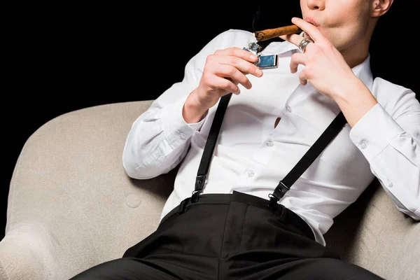 Cropped view of man holding lighter near cigar while sitting in armchair isolated on black — Stock Photo