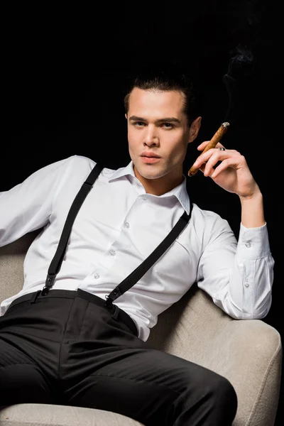 Bel homme tenant cigare assis dans un fauteuil isolé sur noir — Photo de stock