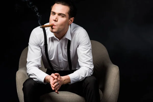 Hombre guapo fumar cigarro mientras está sentado en el sillón con las manos apretadas aisladas en negro - foto de stock