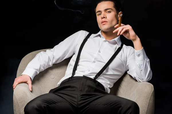 Серйозний чоловік тримає сигару, сидячи в кріслі ізольовано на чорному — стокове фото