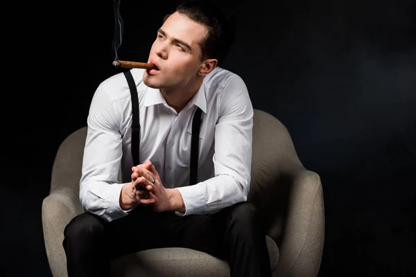 Hombre serio fumar cigarro mientras está sentado en el sillón con las manos apretadas aisladas en negro - foto de stock