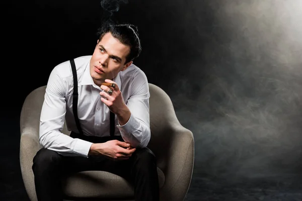 Serious man holding cigar while sitting in armchair on black with smoke — Stock Photo