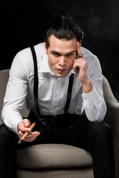 Hombre serio sosteniendo cigarro mientras está sentado en sillón y tocando templo aislado en negro - foto de stock