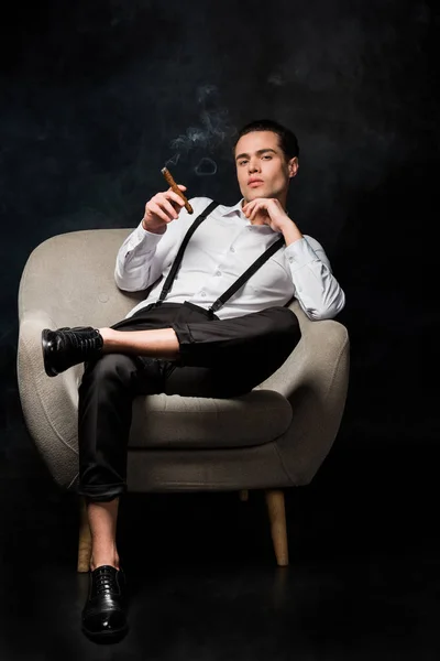 Confident man holding cigar while sitting in armchair on black with smoke — Stock Photo