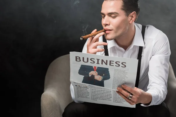 Hombre sentado en sillón leyendo periódico de negocios y fumando cigarro en negro con humo - foto de stock