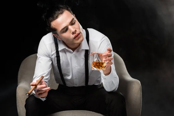 Hombre guapo sentado en sillón mirando un vaso de whisky y sosteniendo cigarro en negro - foto de stock